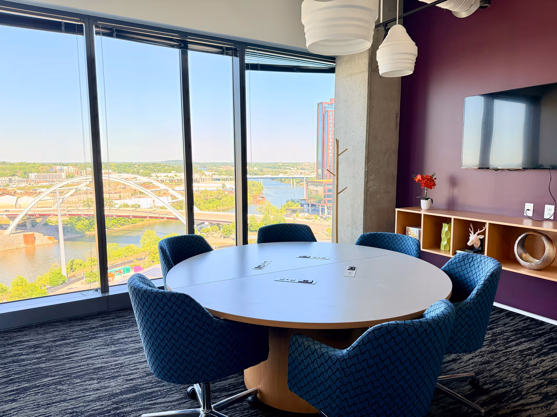 Round white table surrounded by blue chairs in a bright interior with large river view windows.