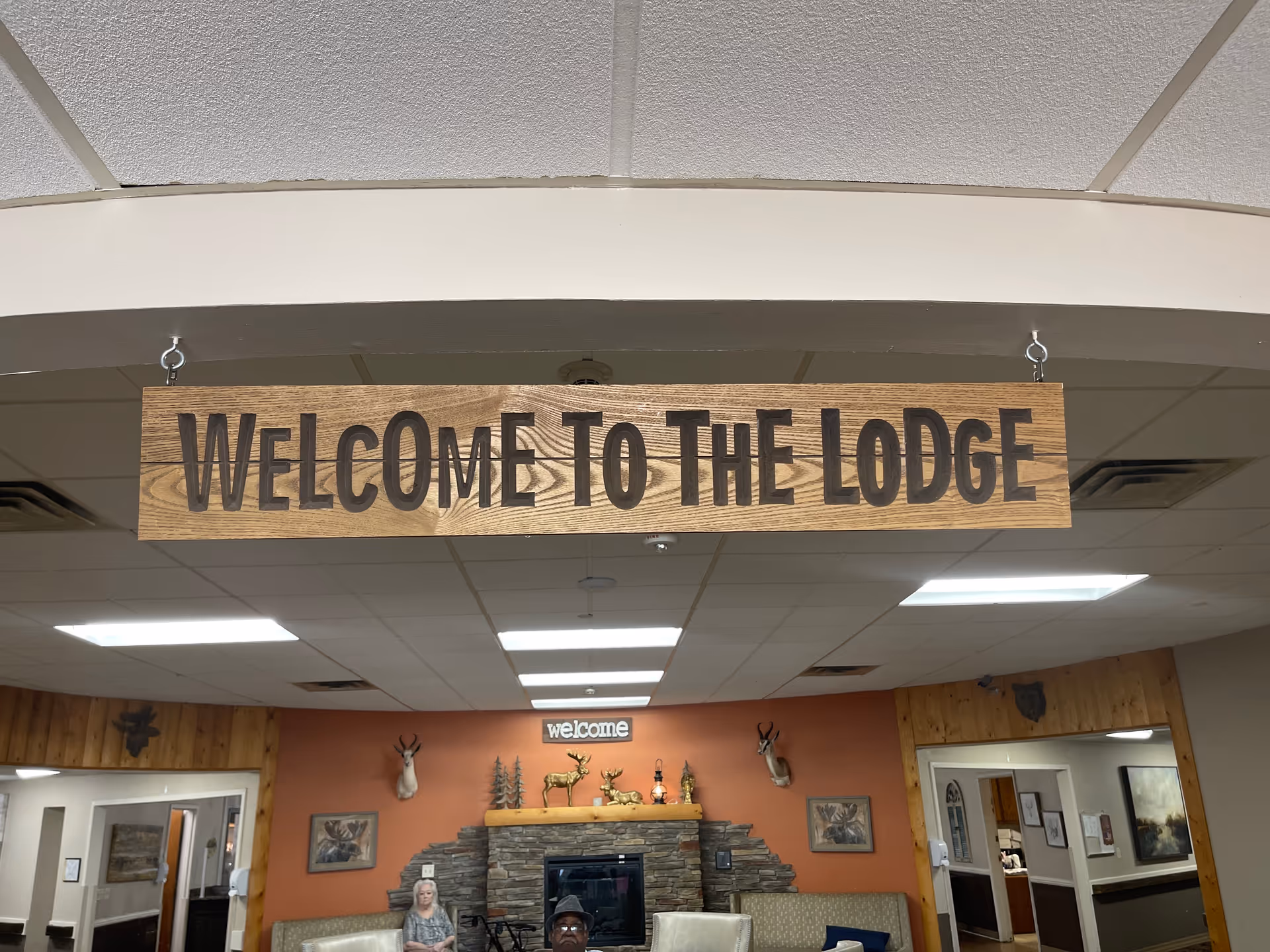 Wooden sign reading 'Welcome to The Lodge' hangs above a cozy lobby with a fireplace.