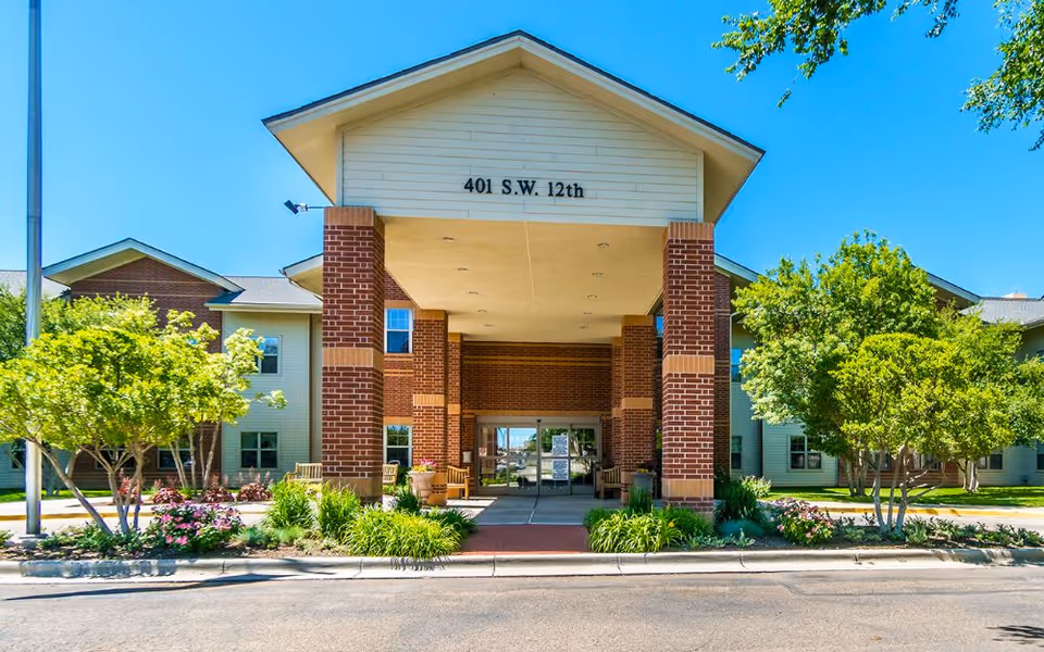 Front exterior view of a senior living facility building with a covered entrance supported by brick columns, surrounded by green trees and landscaping under a clear blue sky. The address 401 S.W. 12th is displayed above the entrance.