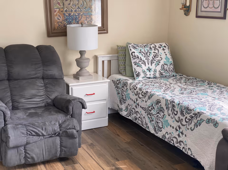 Cozy bedroom with a single bed, patterned quilt, nightstand, lamp, and a recliner.