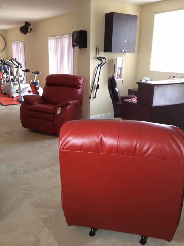 Interior view of a senior living facility lounge area with two red leather recliners in the foreground and exercise equipment, including stationary bikes, visible in the background near windows with vertical blinds. A small reception desk and a brown office chair are also visible near a wall with mounted cabinets and walking canes hanging.