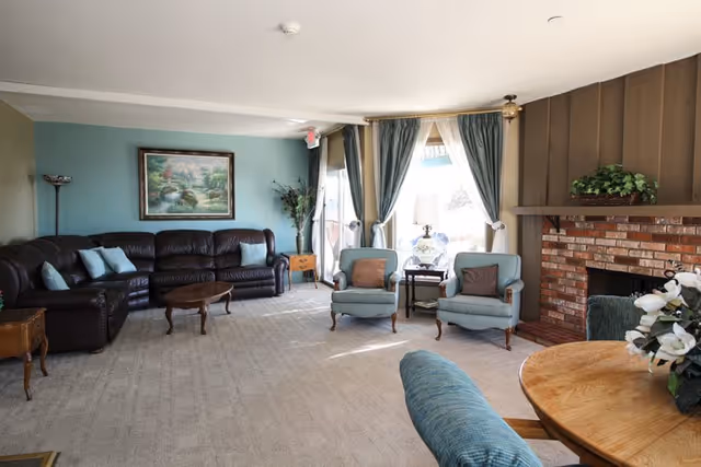 A spacious living room with a large dark brown leather sectional sofa, two light blue armchairs with brown cushions, a wooden coffee table, and side tables. The room features a brick fireplace with a plant on the mantle, large windows with blue curtains letting in natural light, a floor lamp, and a framed landscape painting on a light blue wall.