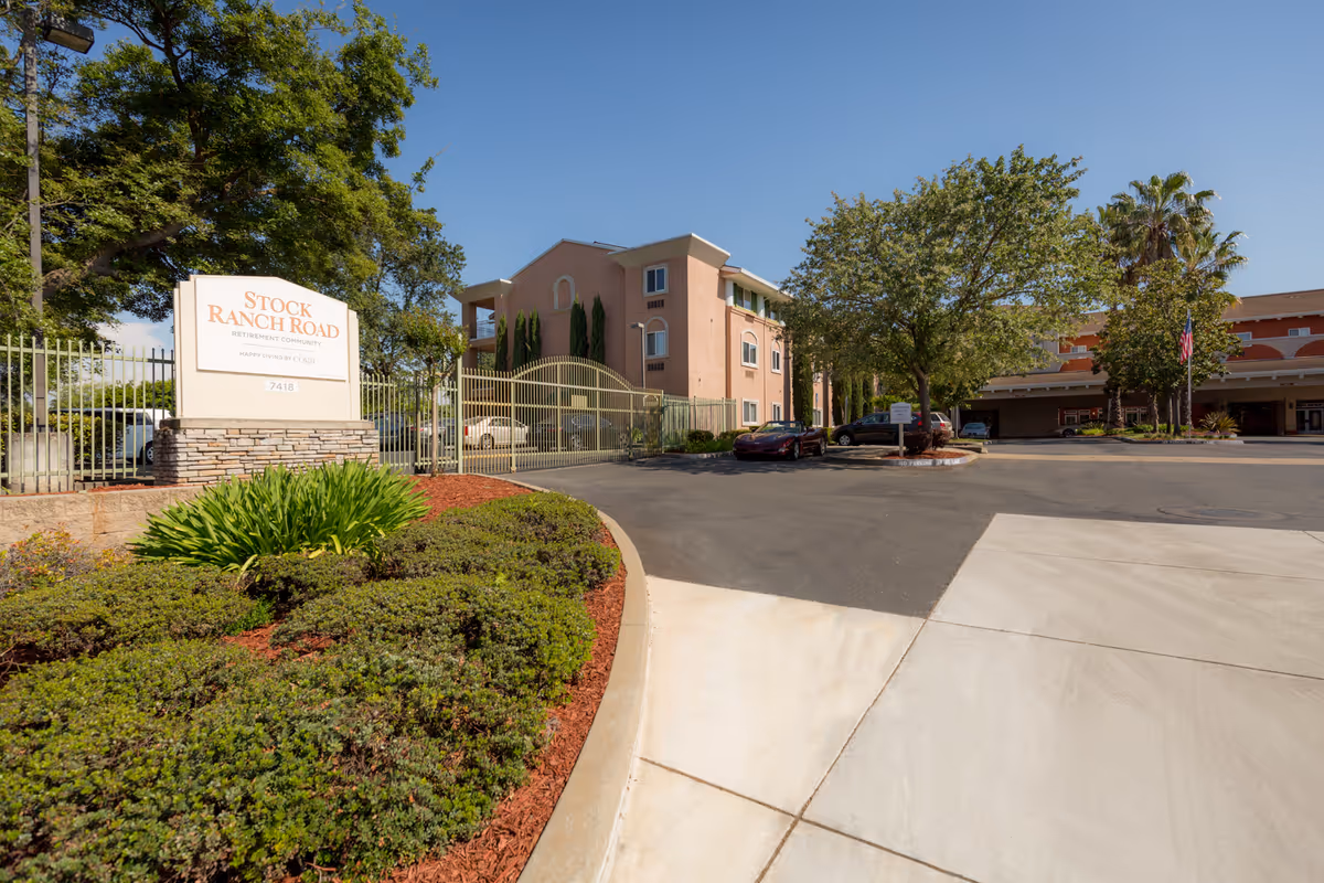Exterior view of Stock Ranch Road Retirement Community showing the entrance sign, gated parking area, and a multi-story building surrounded by trees and landscaping under a clear blue sky.