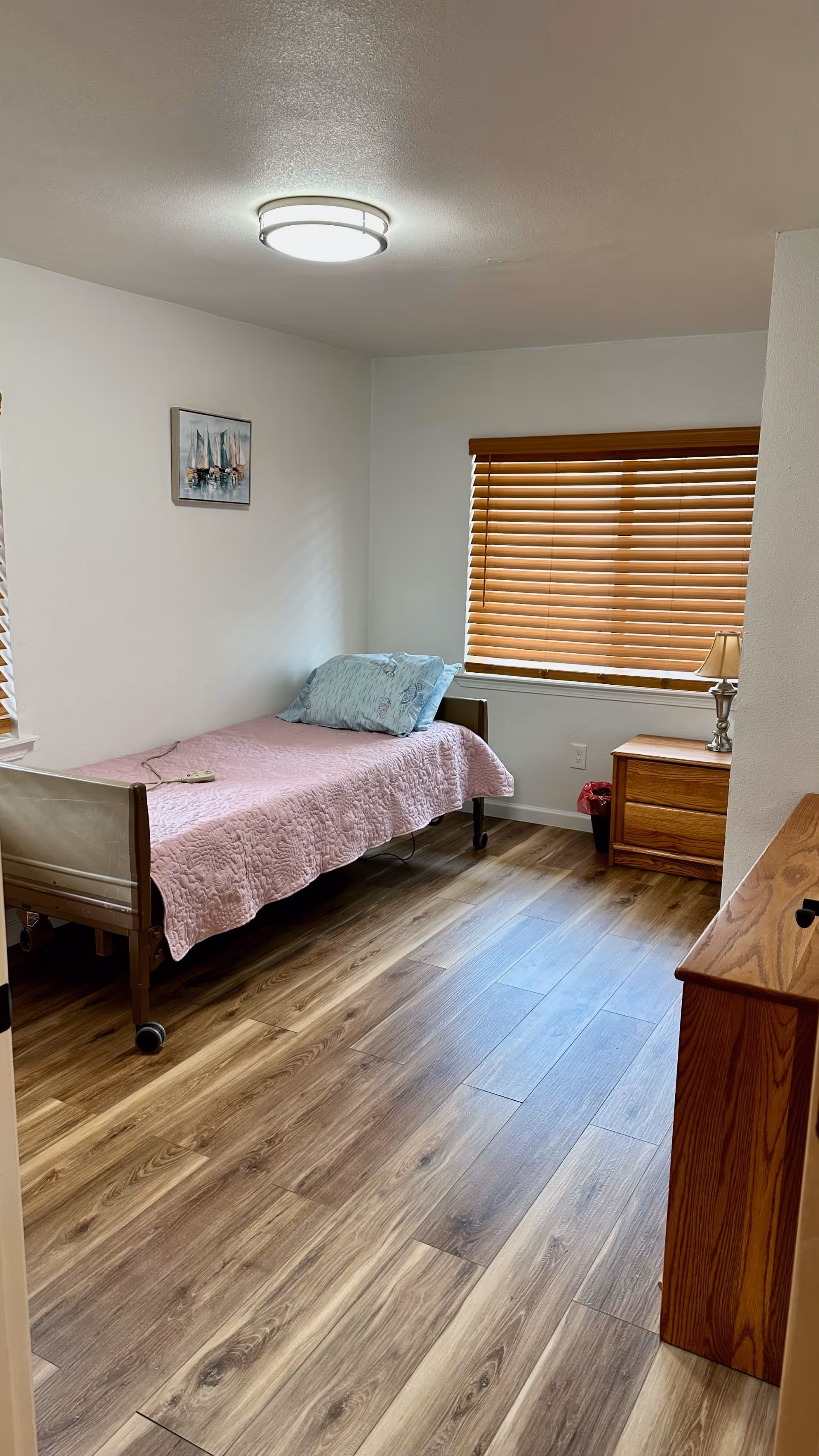 A simple bedroom with a single bed covered with a pink quilt and a blue pillow. There is a wooden nightstand with a lamp next to the window with closed wooden blinds. The floor is wood laminate, and a small painting of sailboats hangs on the wall.