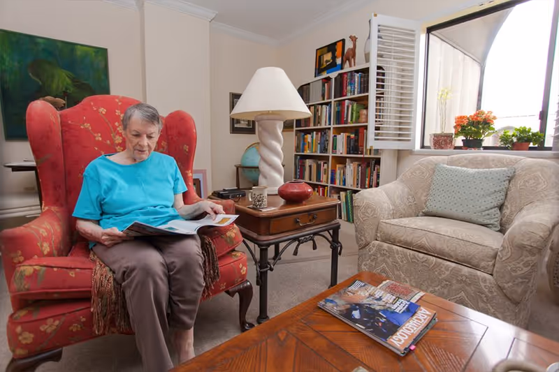 An elderly woman wearing a turquoise shirt and brown pants is sitting in a red floral armchair reading a magazine in a cozy living room. The room features a beige patterned armchair with a light blue pillow, a wooden coffee table with magazines, a side table with a lamp and decorative items, a bookshelf filled with books, and a window with white shutters and potted plants on the windowsill.