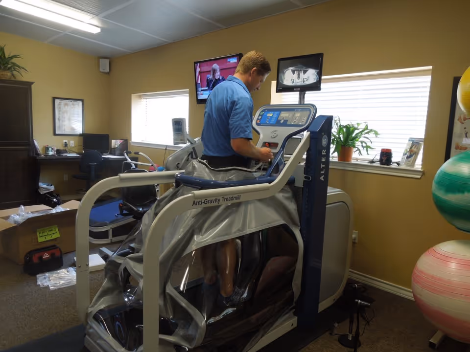 Person on an anti-gravity treadmill in a therapy room with equipment and windows.