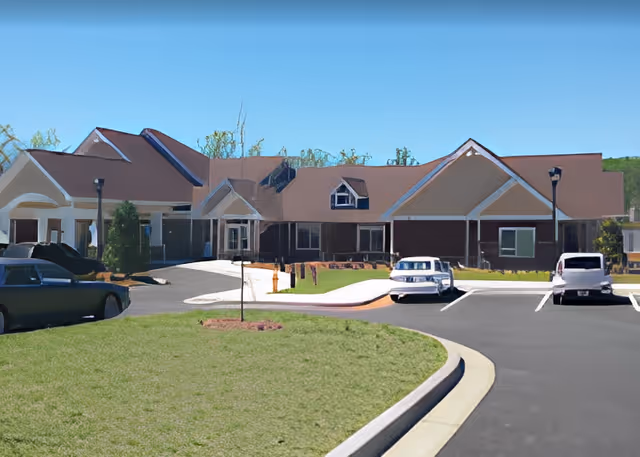 Front exterior of a single-story senior living facility with peaked roofs, an entry canopy, landscaped lawn, and parked cars.