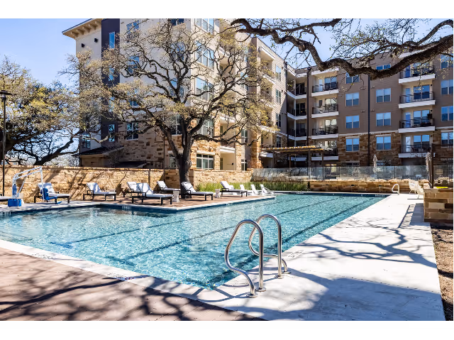 Outdoor swimming pool area with lounge chairs around it, surrounded by a multi-story residential building and large trees casting shadows on the pool deck.
