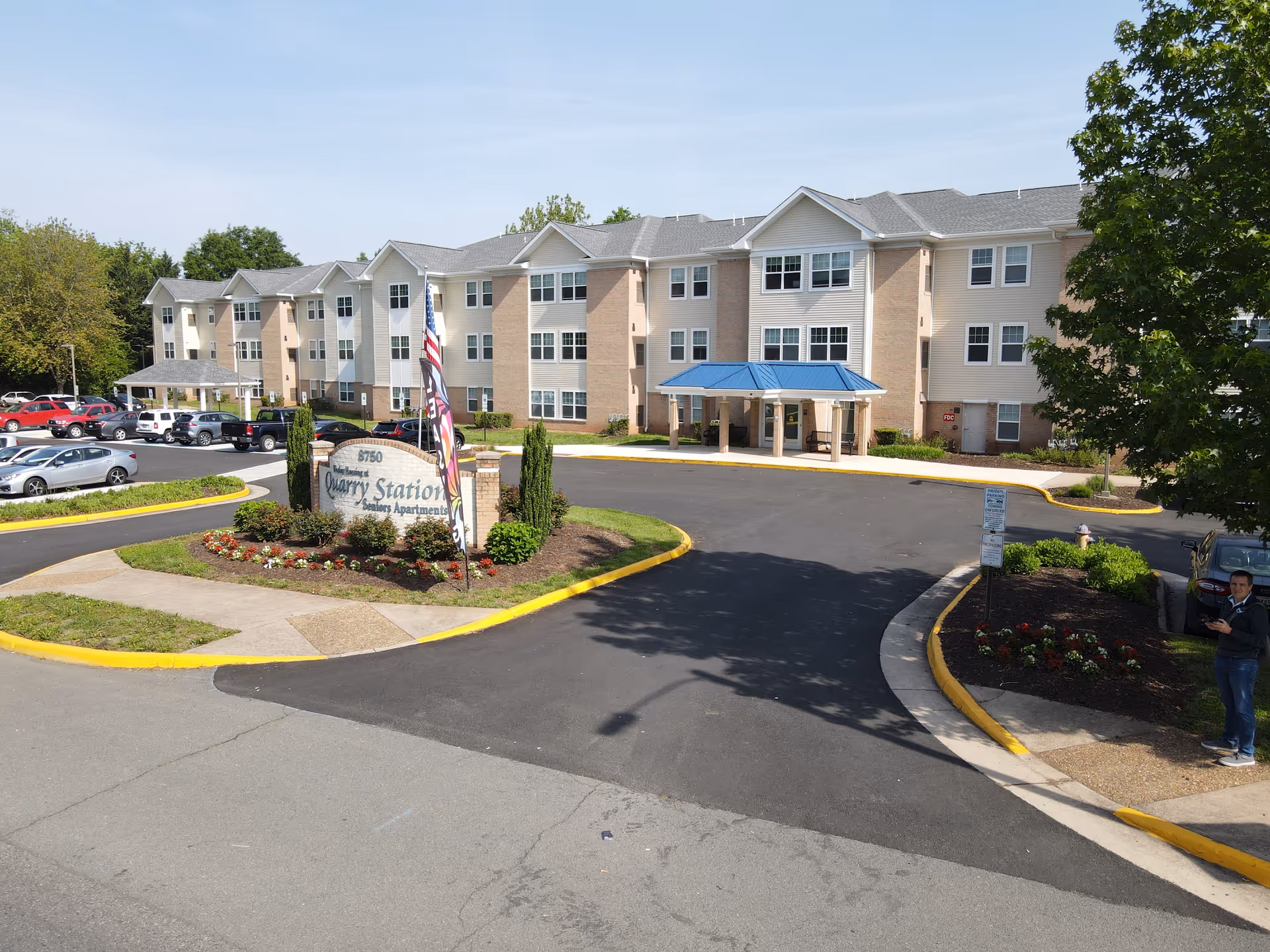 Exterior view of Quarry Station Seniors Apartments, a three-story building with a covered entrance, circular driveway, landscaped sign and parked cars.