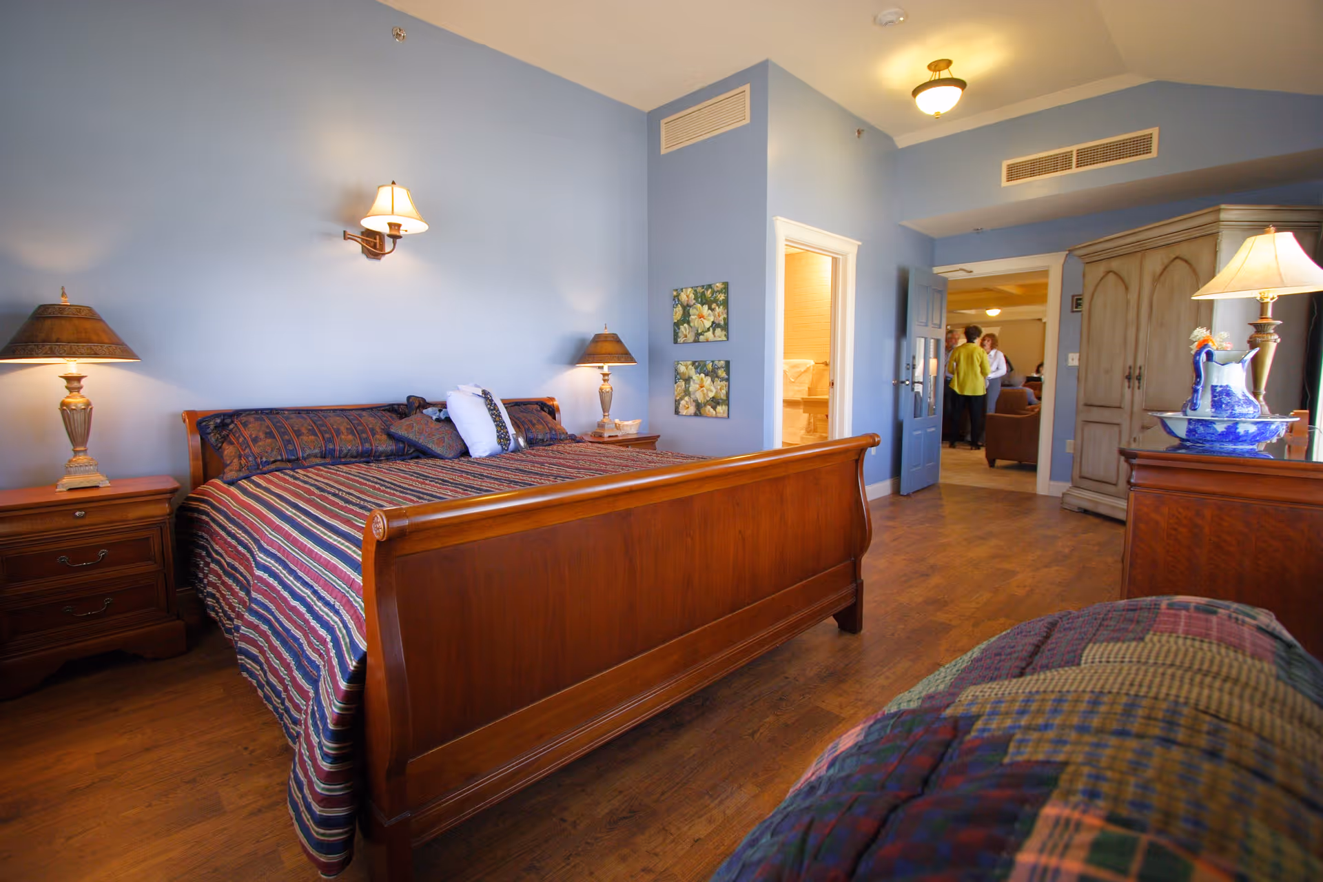 A bright bedroom with a wooden sleigh bed, patterned bedcover, bedside tables and lamps, blue walls, and an open doorway showing people in a hallway.
