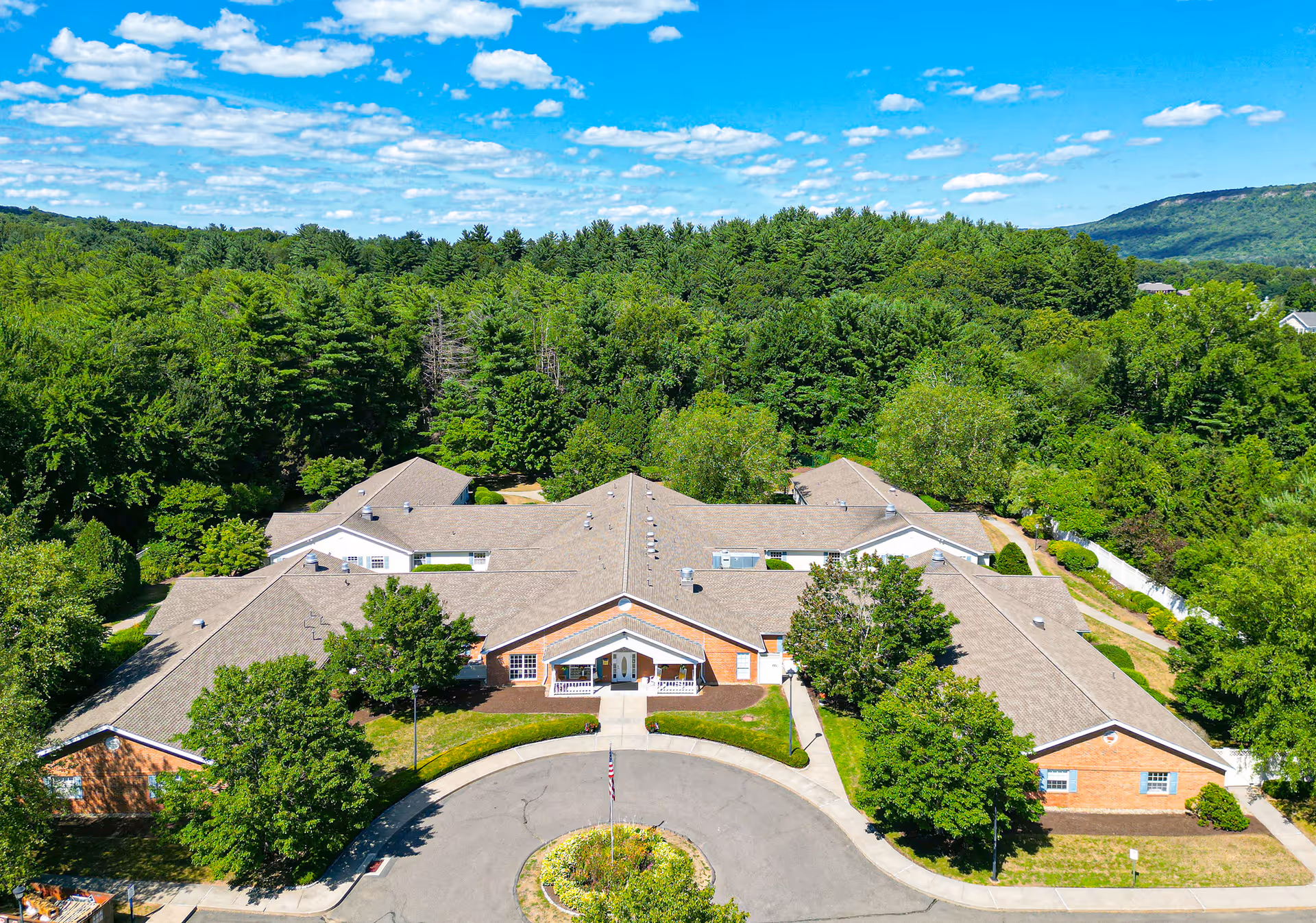 Aerial view of Arden Courts - ProMedica Memory Care Community in Avon, showing a large single-story building with a circular driveway and landscaped greenery surrounded by dense trees under a blue sky with scattered clouds.