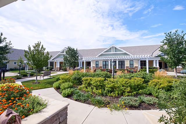 Front exterior of a single-story senior living building with a landscaped courtyard, walkways, benches, and a pergola under a partly cloudy sky.