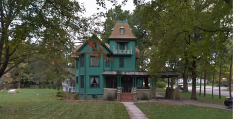 A green turreted house-like building sits on a grassy lawn under tall trees.