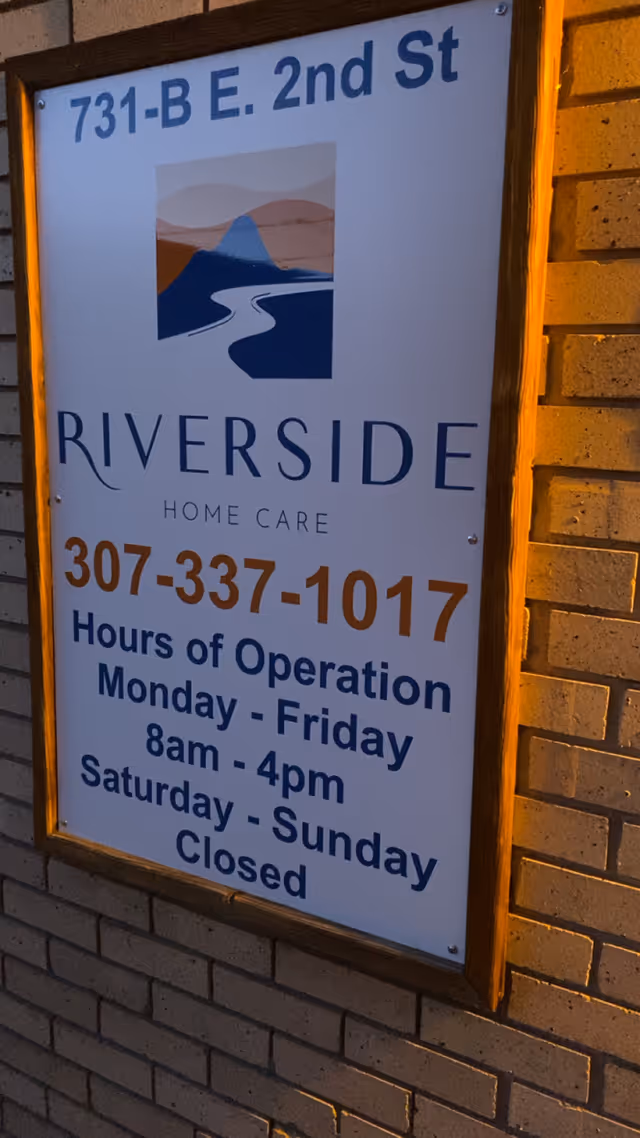 Signboard outside Riverside Home Care on brick wall showing hours and contact info.