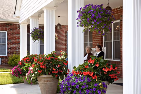 Two residents sit on a flower-filled porch beside a brick building with white columns.