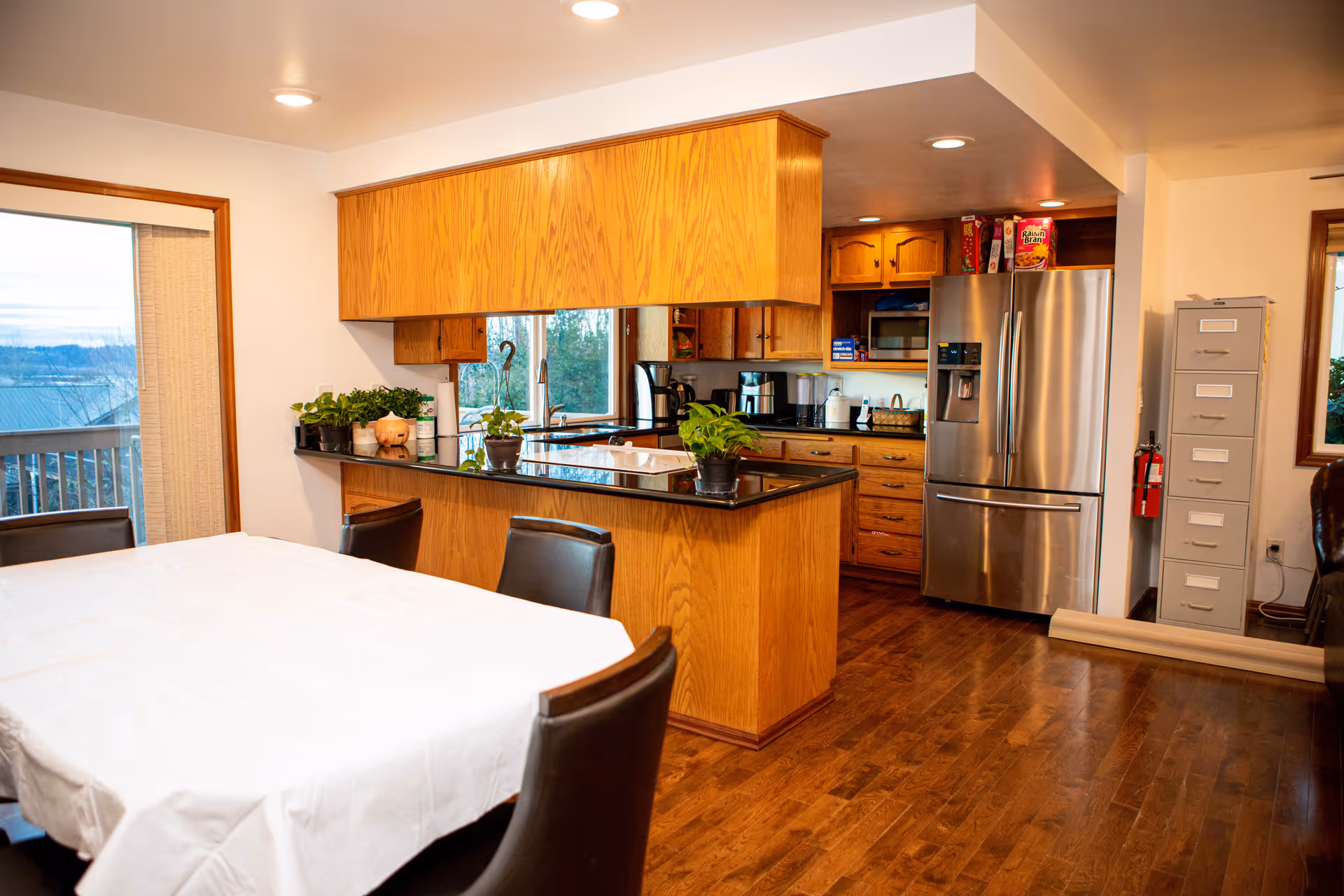 Open-plan kitchen and dining area with wooden cabinets and island, a stainless steel refrigerator, a dining table with a white tablecloth, and potted plants.