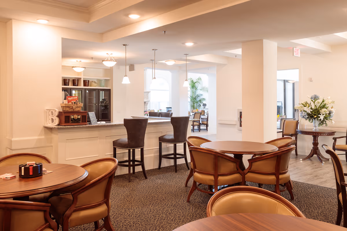 A well-lit common area in a senior living facility featuring round wooden tables with cushioned chairs, a bar counter with two high chairs, pendant lights hanging from the ceiling, and a decorative flower arrangement on a side table. The space has a warm and inviting atmosphere with light-colored walls and carpeted flooring.
