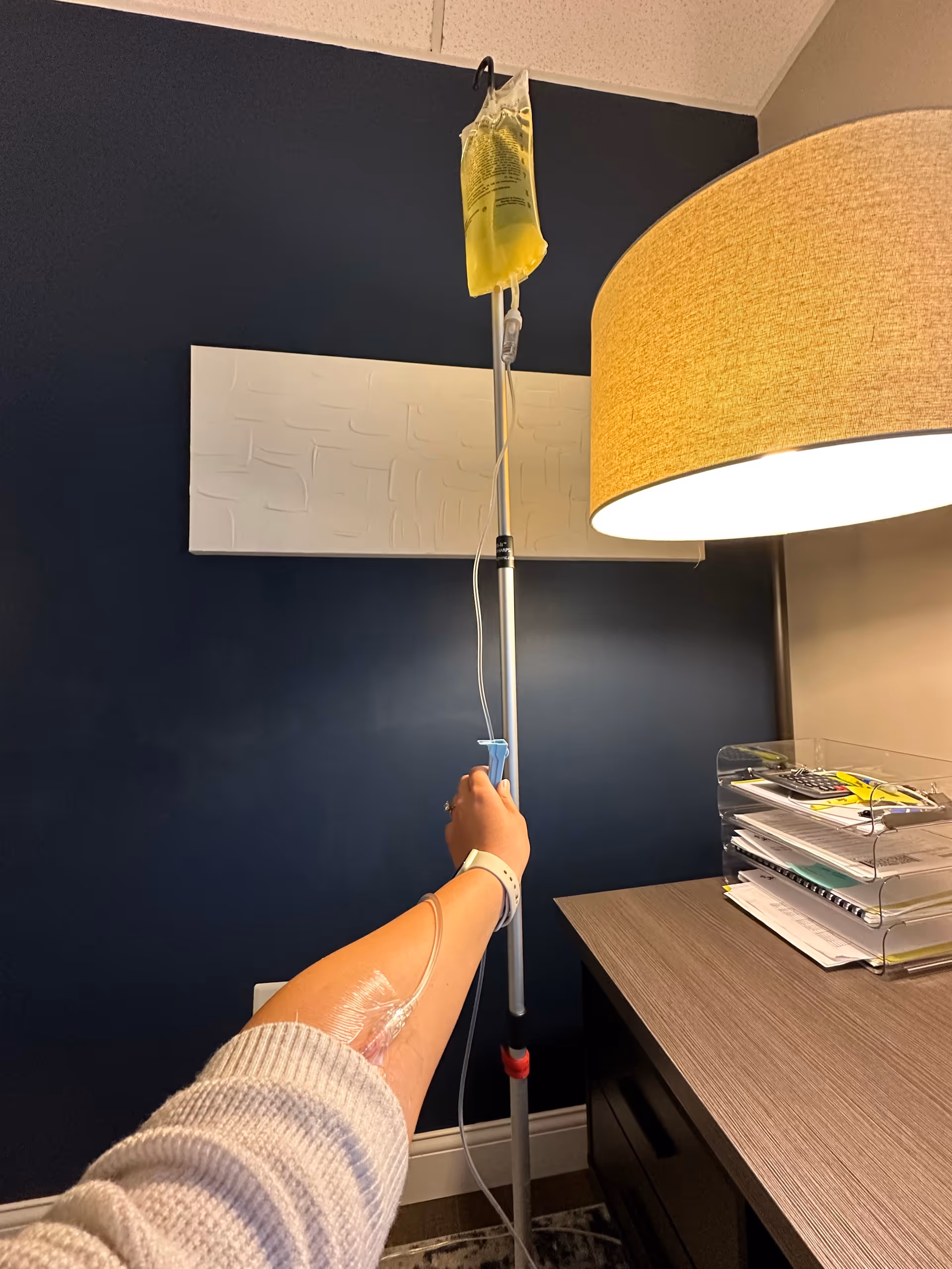 Arm with IV line in a facility room beside a desk and lamp.