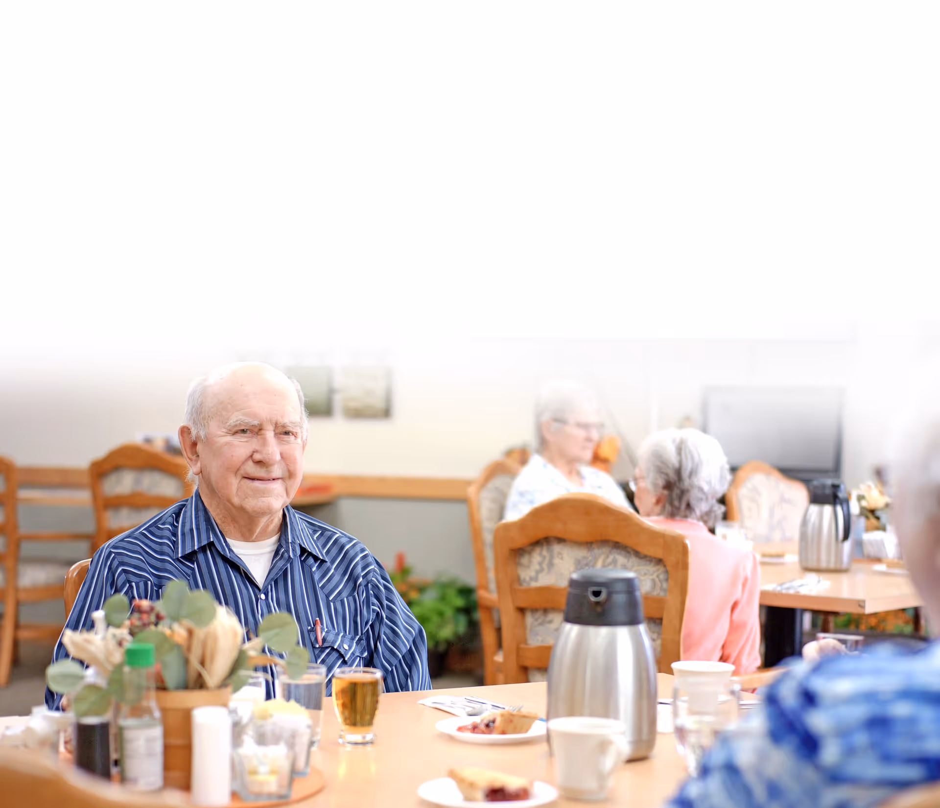 An elderly man sits at a dining table in a senior living facility dining room with other residents and tableware on the table.