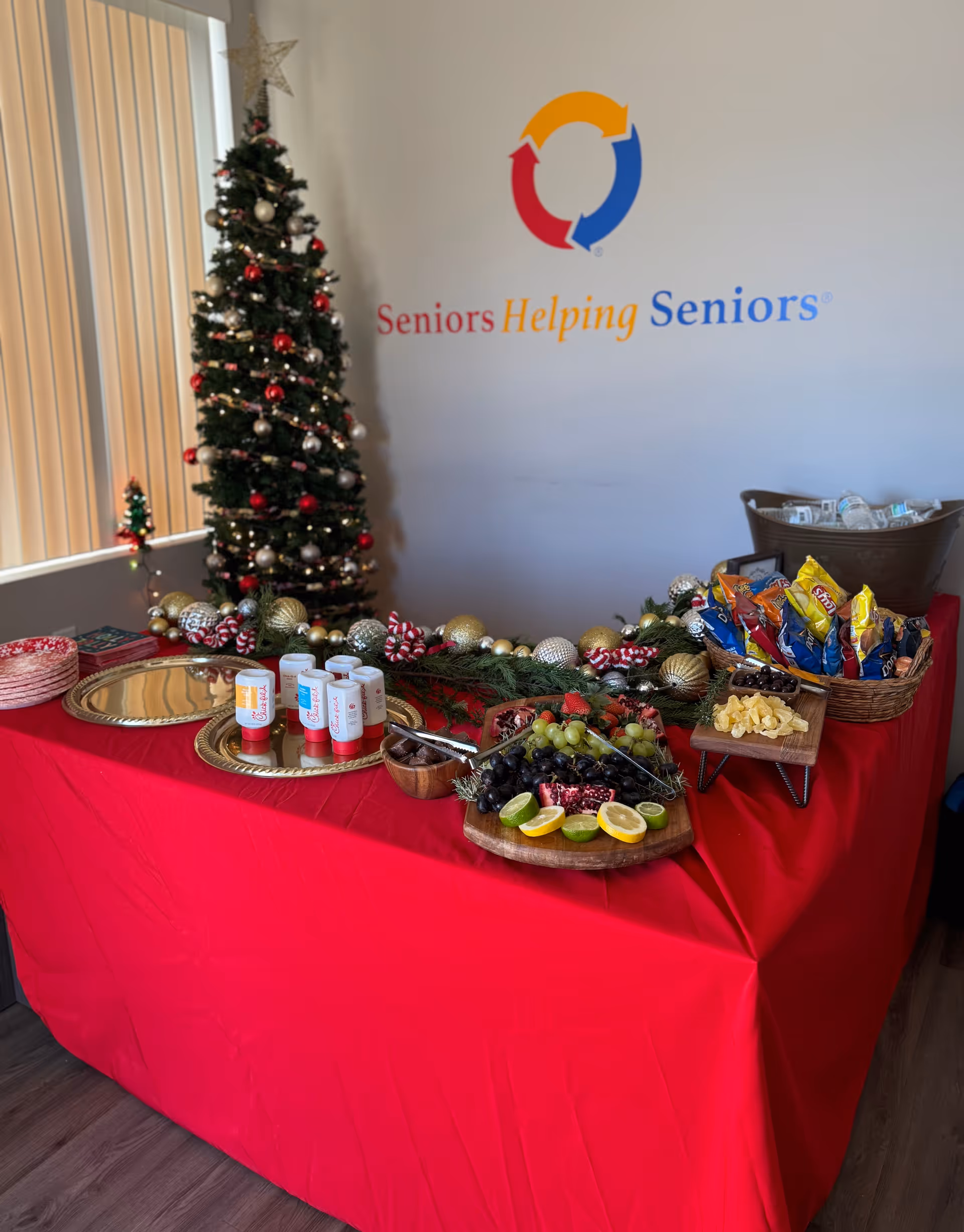 Indoor display table with red cloth, fruit, chips, and a Christmas tree beside logo.