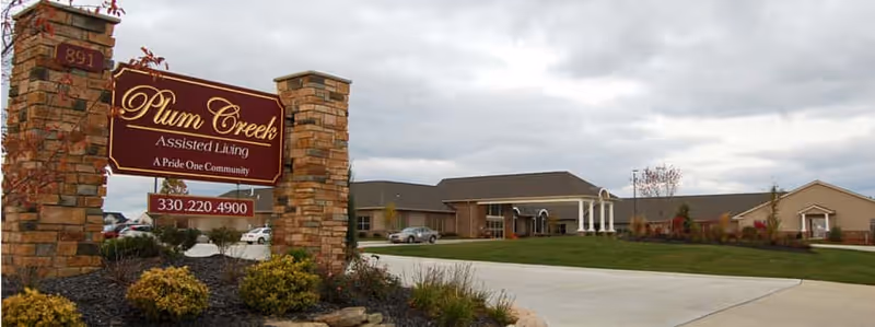 Stone entrance sign reading "Plum Creek Assisted Living" in front of a single-story assisted living building, driveway, and landscaped lawn under a cloudy sky.