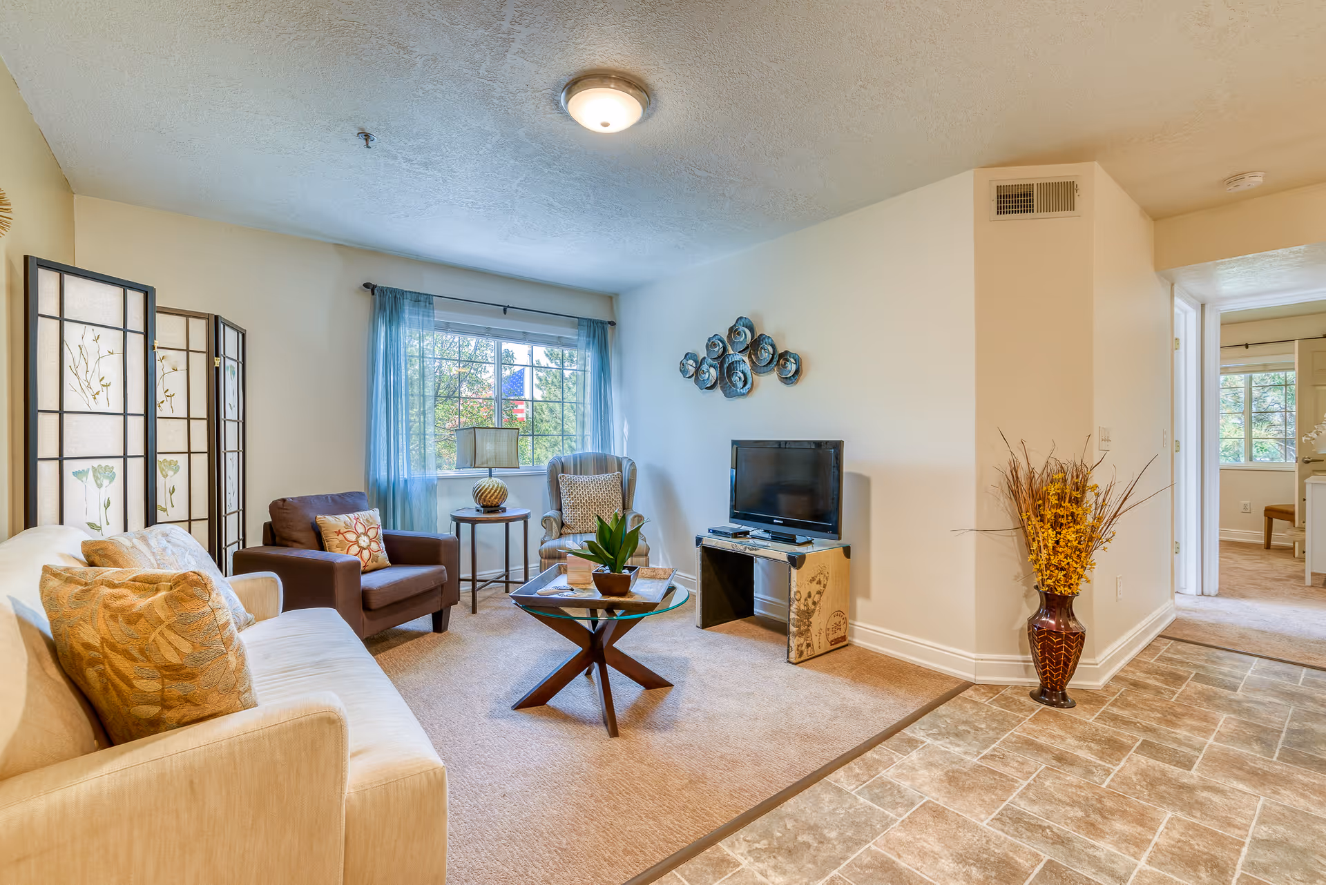 Bright living room in a senior facility with sofa, chairs, TV, and decorative folding screen.