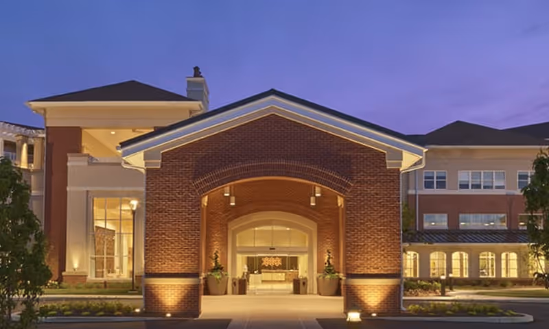 Front exterior view of Woodcrest Villa by Tandem Living at dusk, featuring a large brick entrance with an arched doorway, illuminated by warm lighting, with the main building visible in the background.