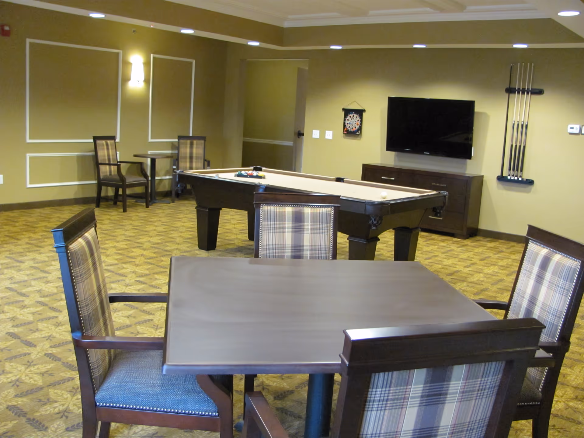 Interior community game room with a pool table, wall-mounted TV, and a table surrounded by plaid-upholstered chairs.
