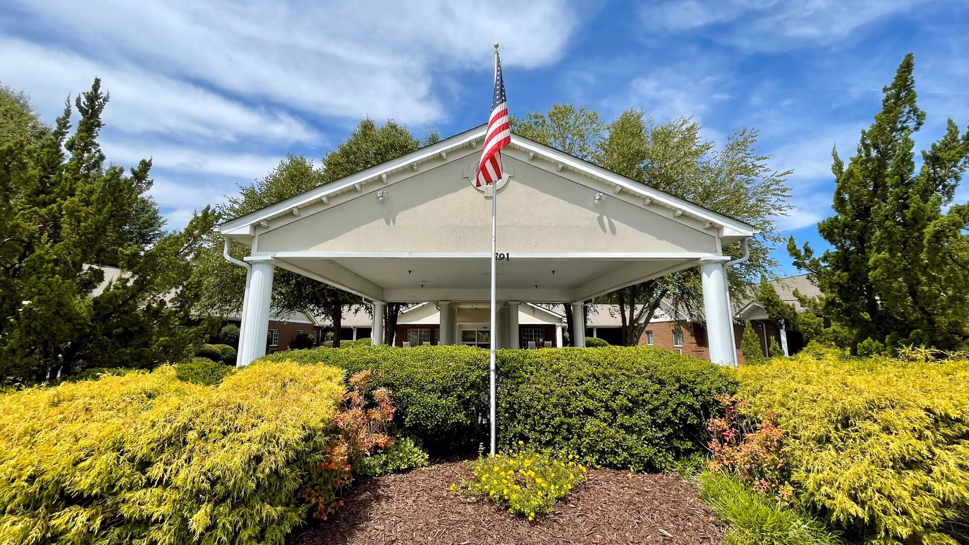 Covered front entrance of a nursing facility with an American flag and manicured shrubs.