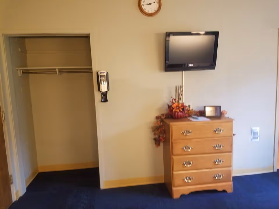 A room corner with a wooden chest of drawers decorated with autumn-themed items, a wall-mounted flat screen TV above it, a clock on the wall, an open closet with a hanging rod, and a wall-mounted hand sanitizer dispenser. The floor is carpeted in blue.