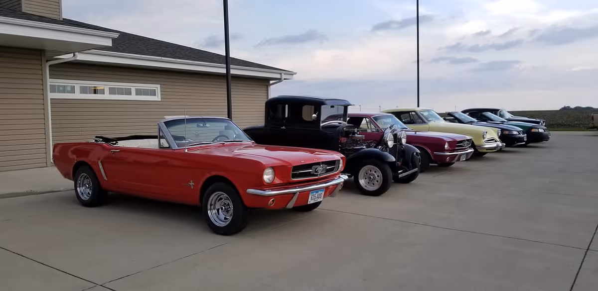 A row of vintage and classic cars parked on a concrete driveway outside a beige building under a cloudy sky.