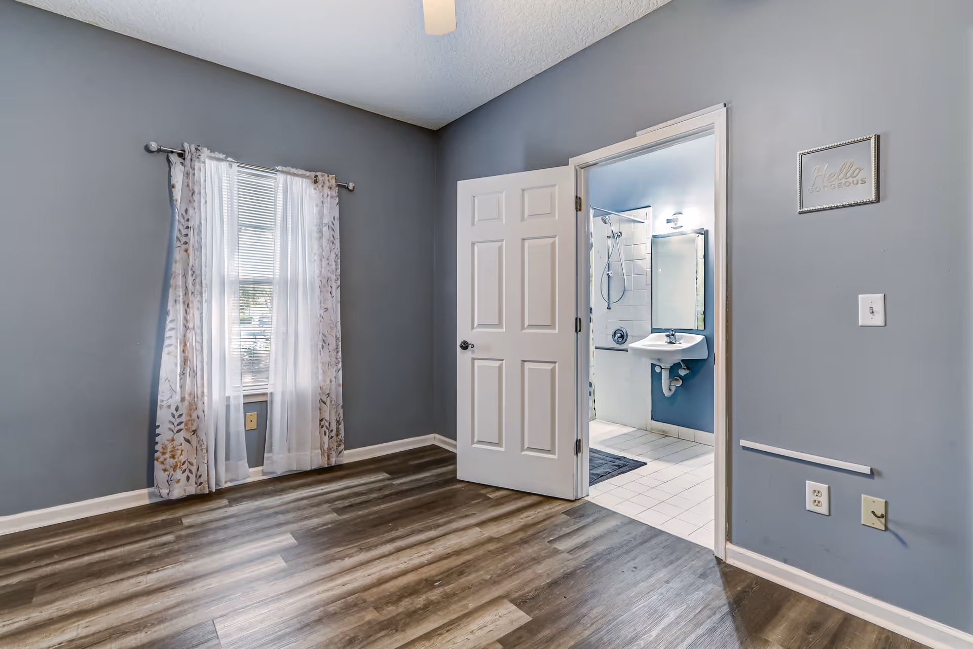 Blue-walled room with window and floral curtains beside a doorway to a bathroom.