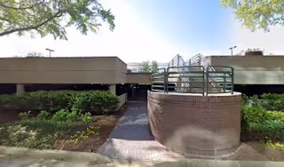 Exterior view of a low-rise facility entrance with brick curved wall, railing, and surrounding landscaping.