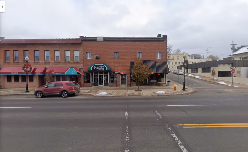 A street view of brick storefronts and a parked SUV along a downtown road.