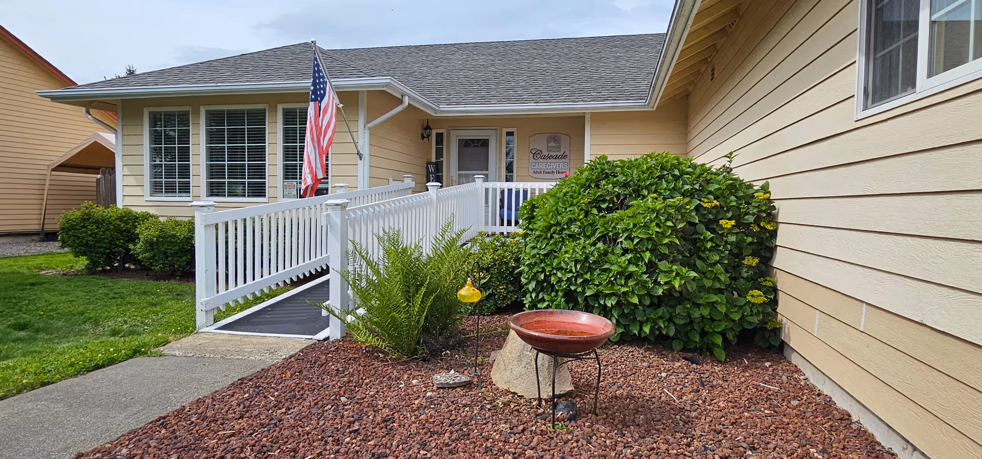 Front exterior of a single-story yellow adult family home with a white wheelchair ramp, American flag, and landscaped front yard.