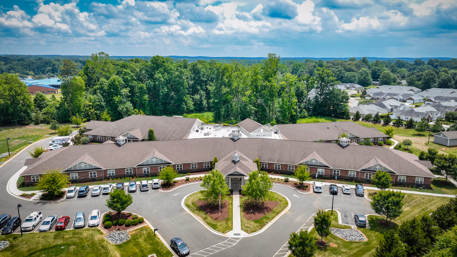 Aerial view of Mebane Ridge Assisted Living facility showing a large, single-story building with a brown roof surrounded by parking lots, trees, and landscaped areas. The building is situated in a green, wooded area with residential houses visible in the background under a partly cloudy sky.
