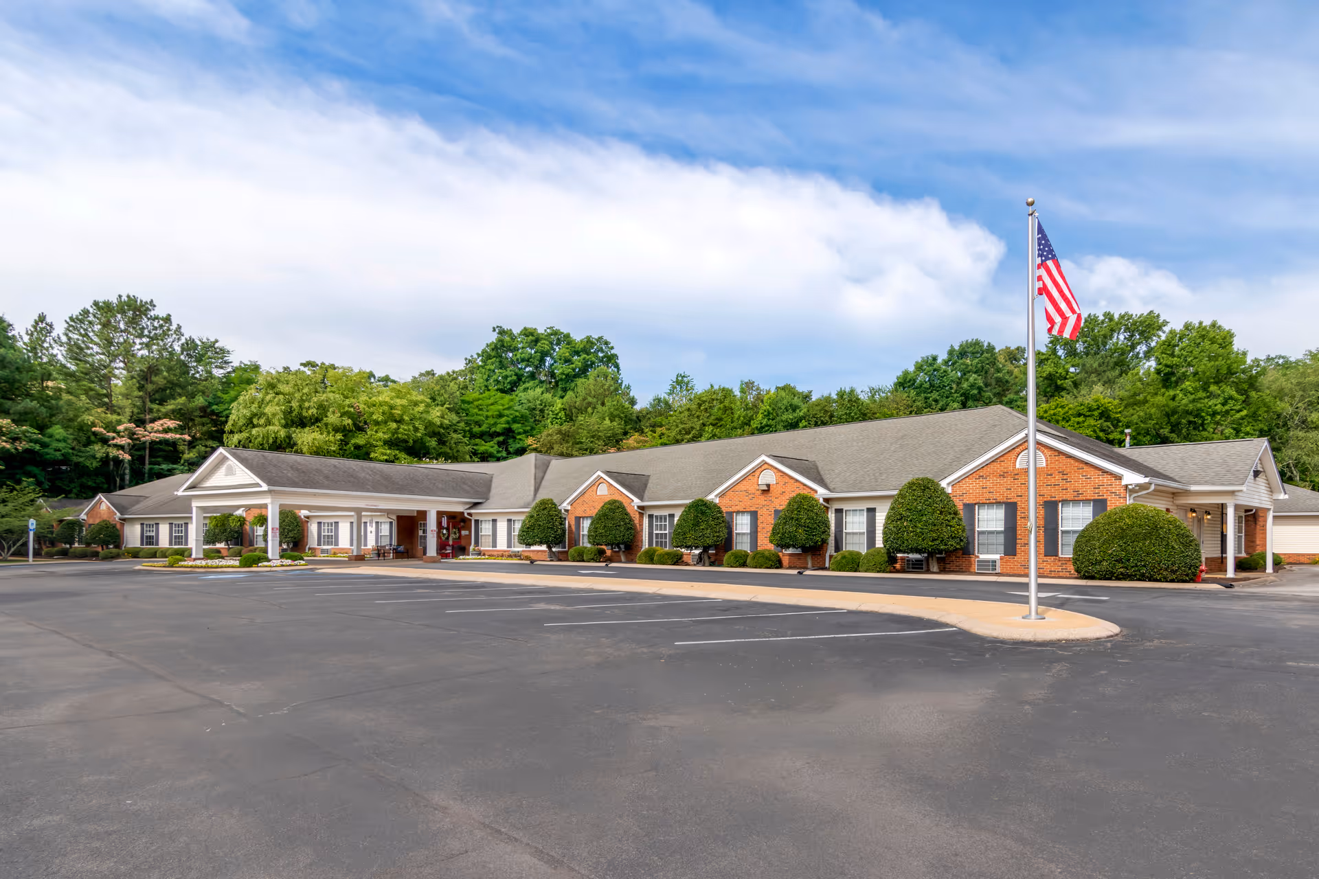 Exterior view of a single-story brick building with white trim, surrounded by neatly trimmed bushes and trees. There is a large paved parking lot in front and an American flag on a flagpole near the entrance. The sky is partly cloudy with blue patches.