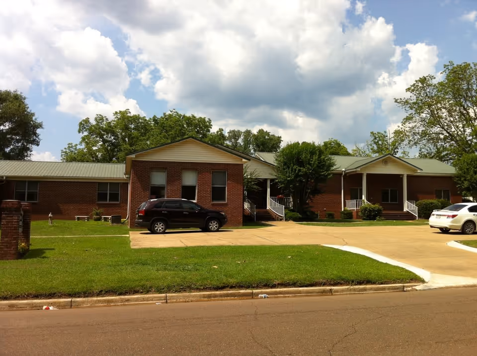 Front view of a single-story brick senior living building with porches, a driveway and parked cars under a partly cloudy sky.
