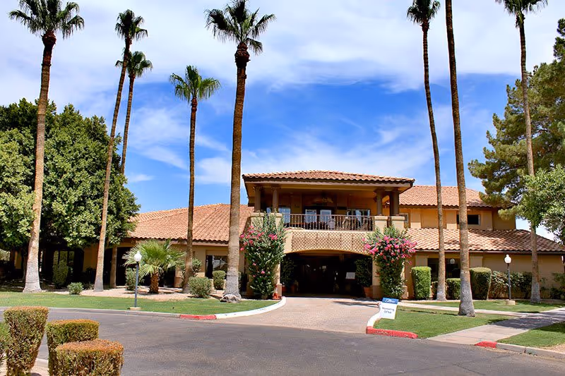 Exterior view of a tan senior living facility with palm trees and a driveway.