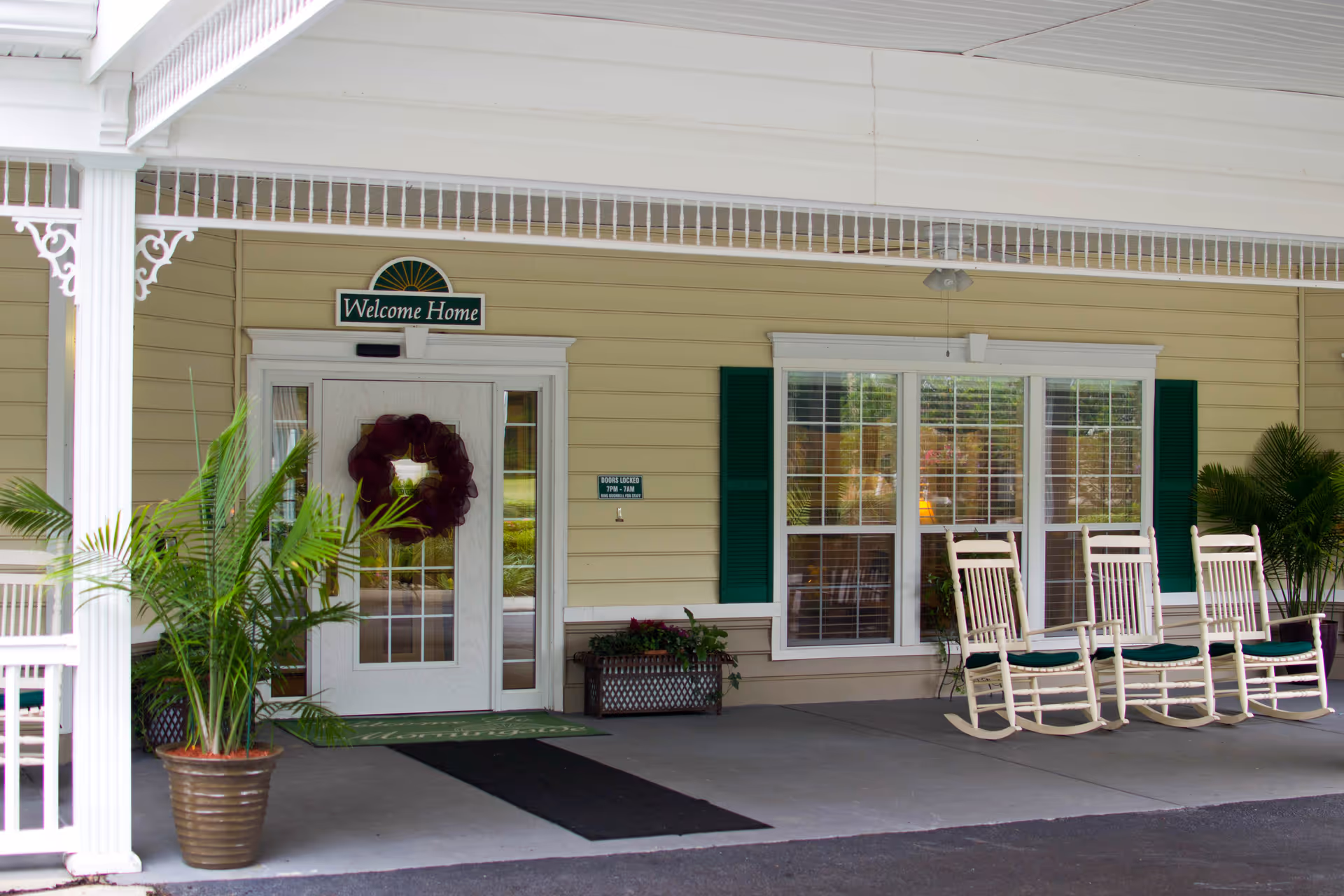 Covered front entrance of a senior living facility with a 'Welcome Home' sign, a wreath on the door, potted plants, and rocking chairs on the porch.