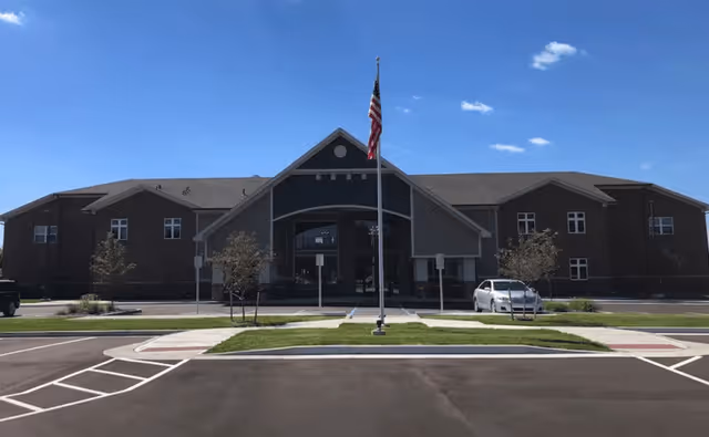 Front exterior of a two-story senior living building with a central covered entrance, American flag on a flagpole, and a parking lot.