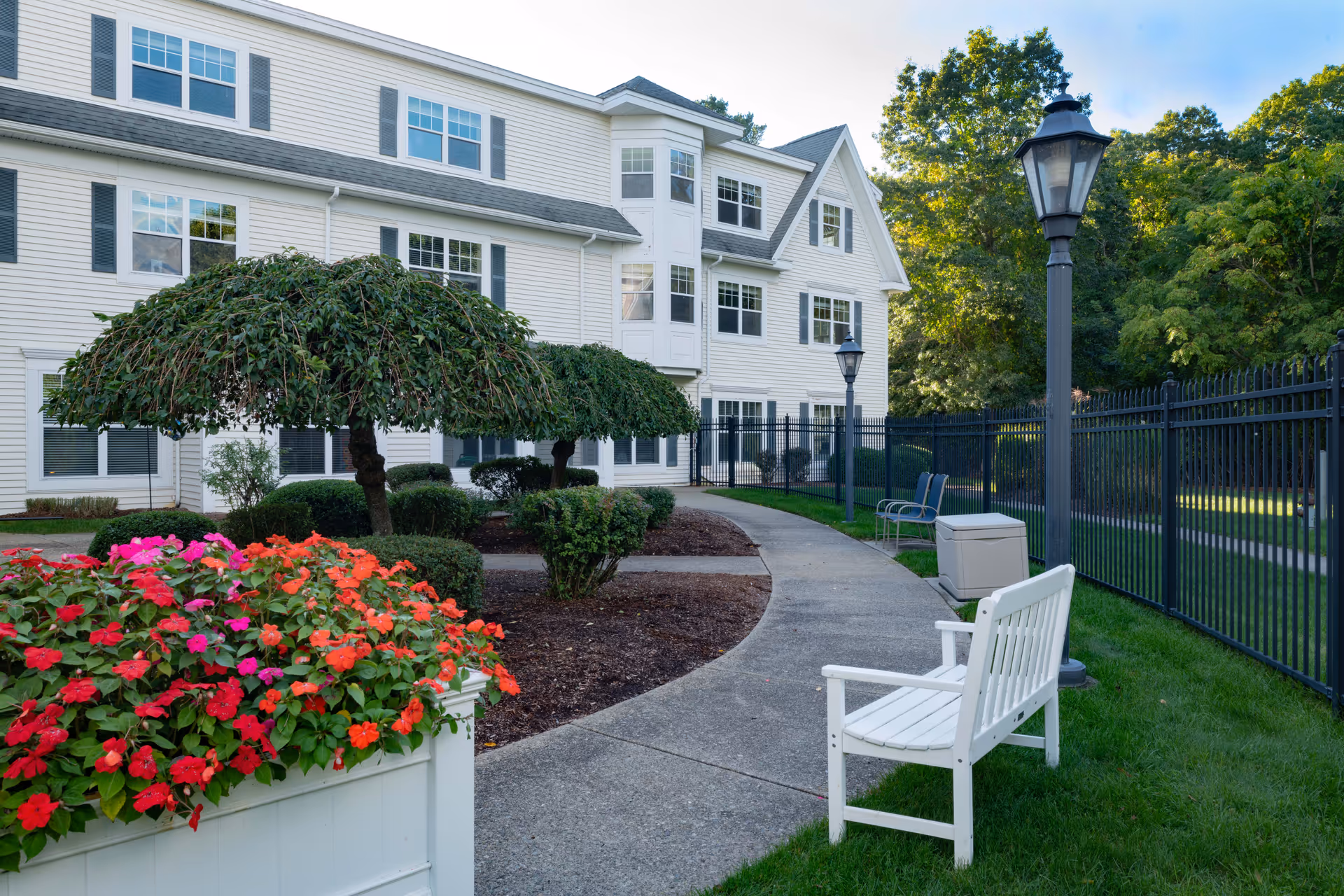 Curved walkway and landscaped courtyard with benches, lamp posts, and flowering planters beside the exterior of a white senior living building.