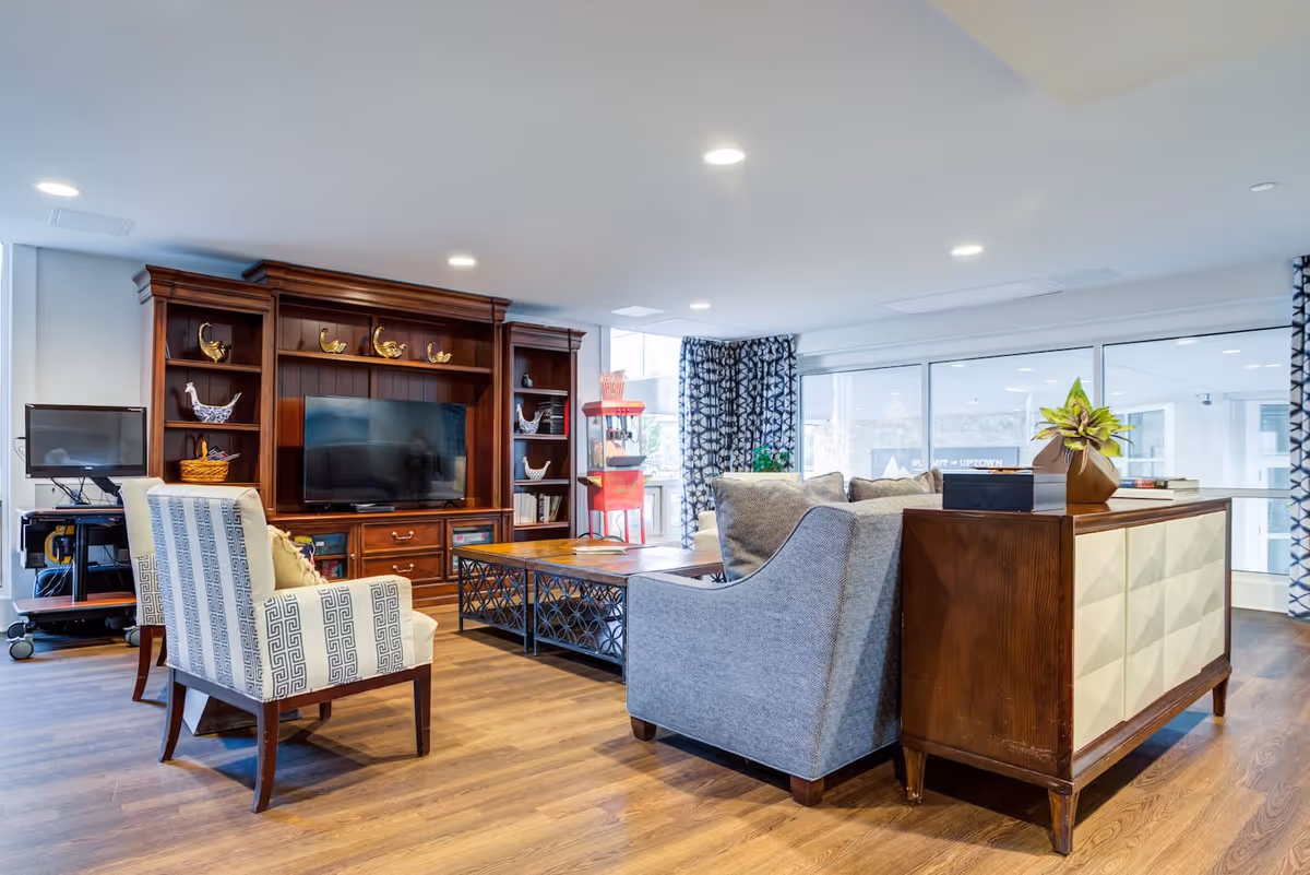 A bright and spacious living room area with wooden flooring, a large wooden entertainment center with a flat-screen TV, two patterned armchairs, a gray sofa, a wooden coffee table with a metal frame, and a sideboard with a plant on top. Large windows with patterned curtains allow natural light to fill the room.