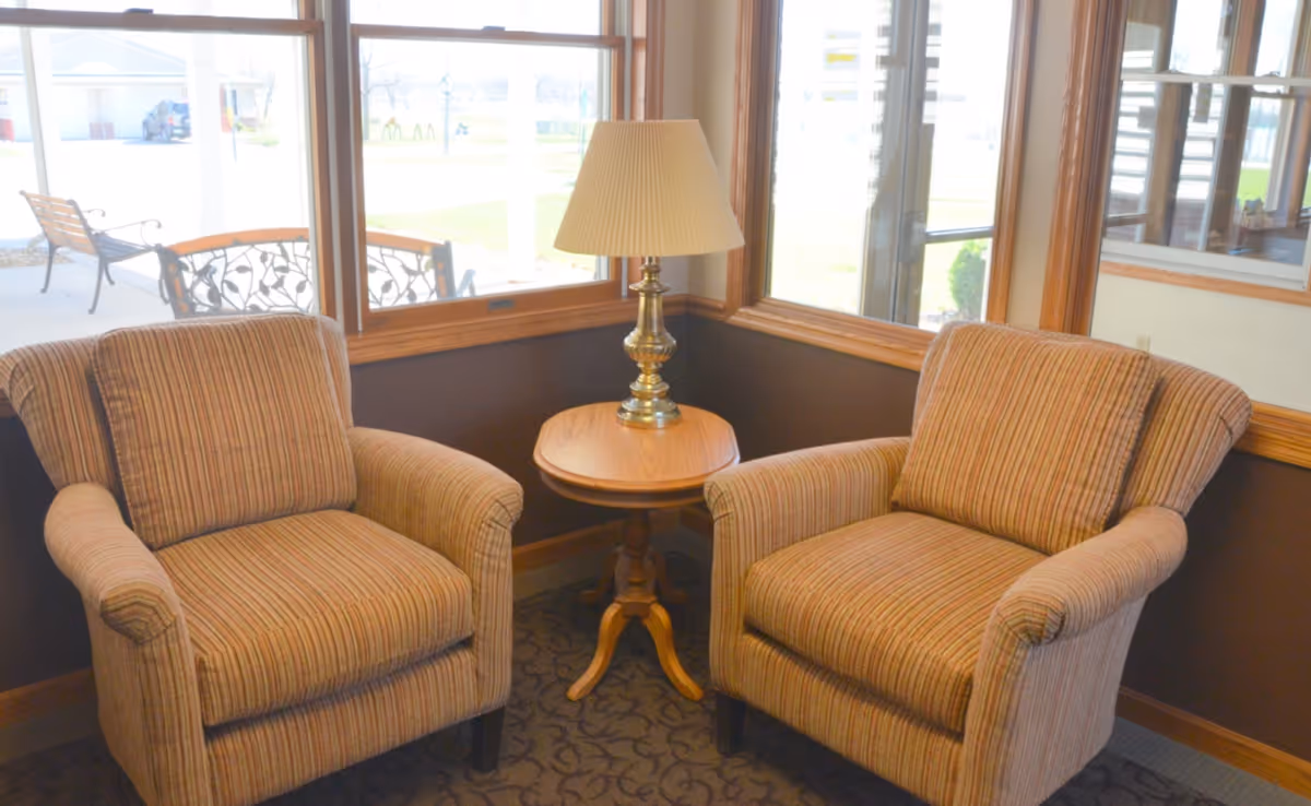 Two upholstered armchairs flanking a wooden side table with a lamp beside windows overlooking an outdoor patio.