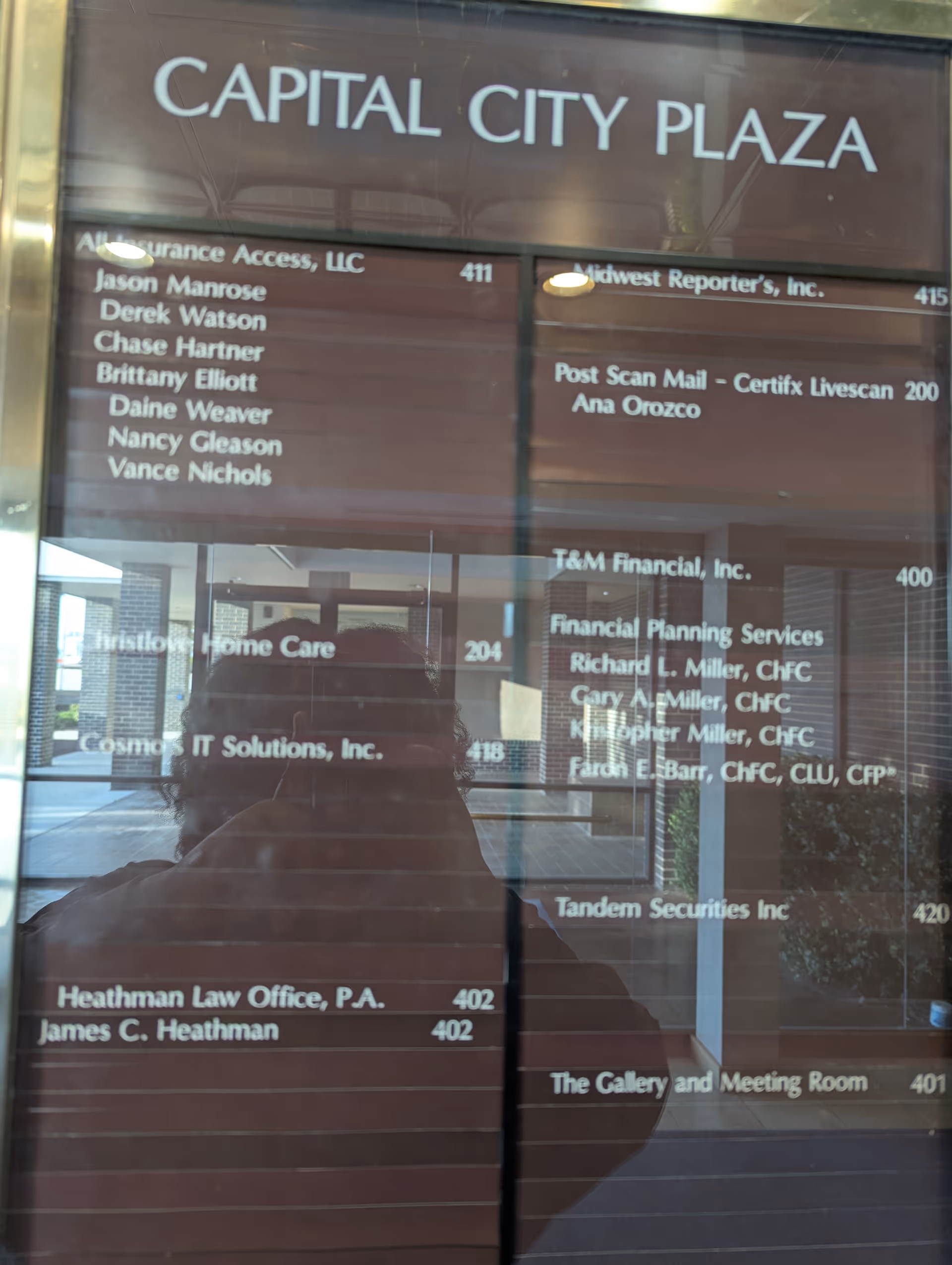 Interior glass directory board at Capital City Plaza with tenant listings; photographer's silhouette reflected.