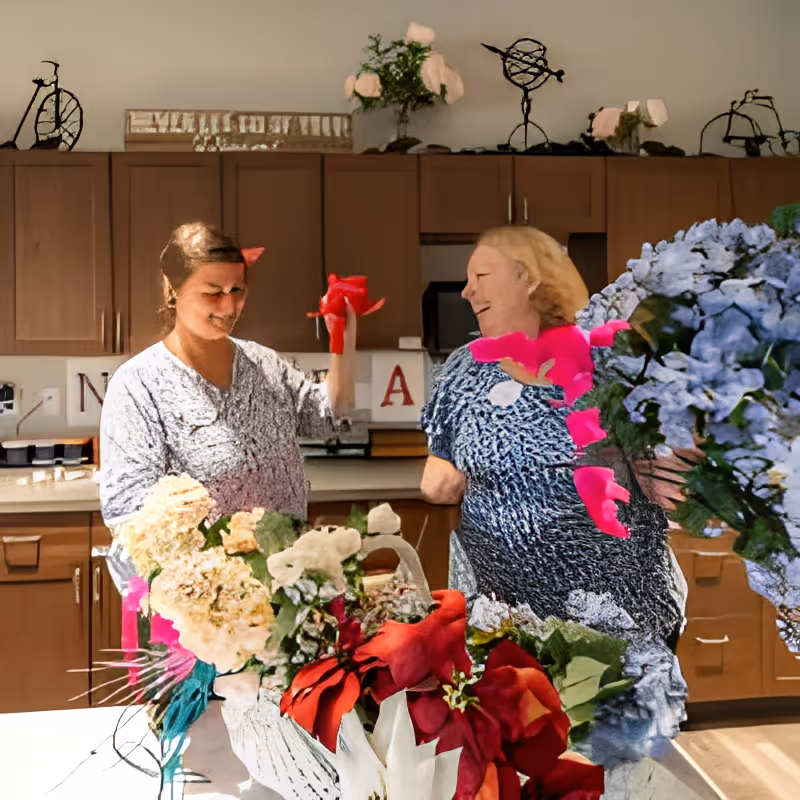 Two women arranging a floral centerpiece on a kitchen counter in a communal living area.