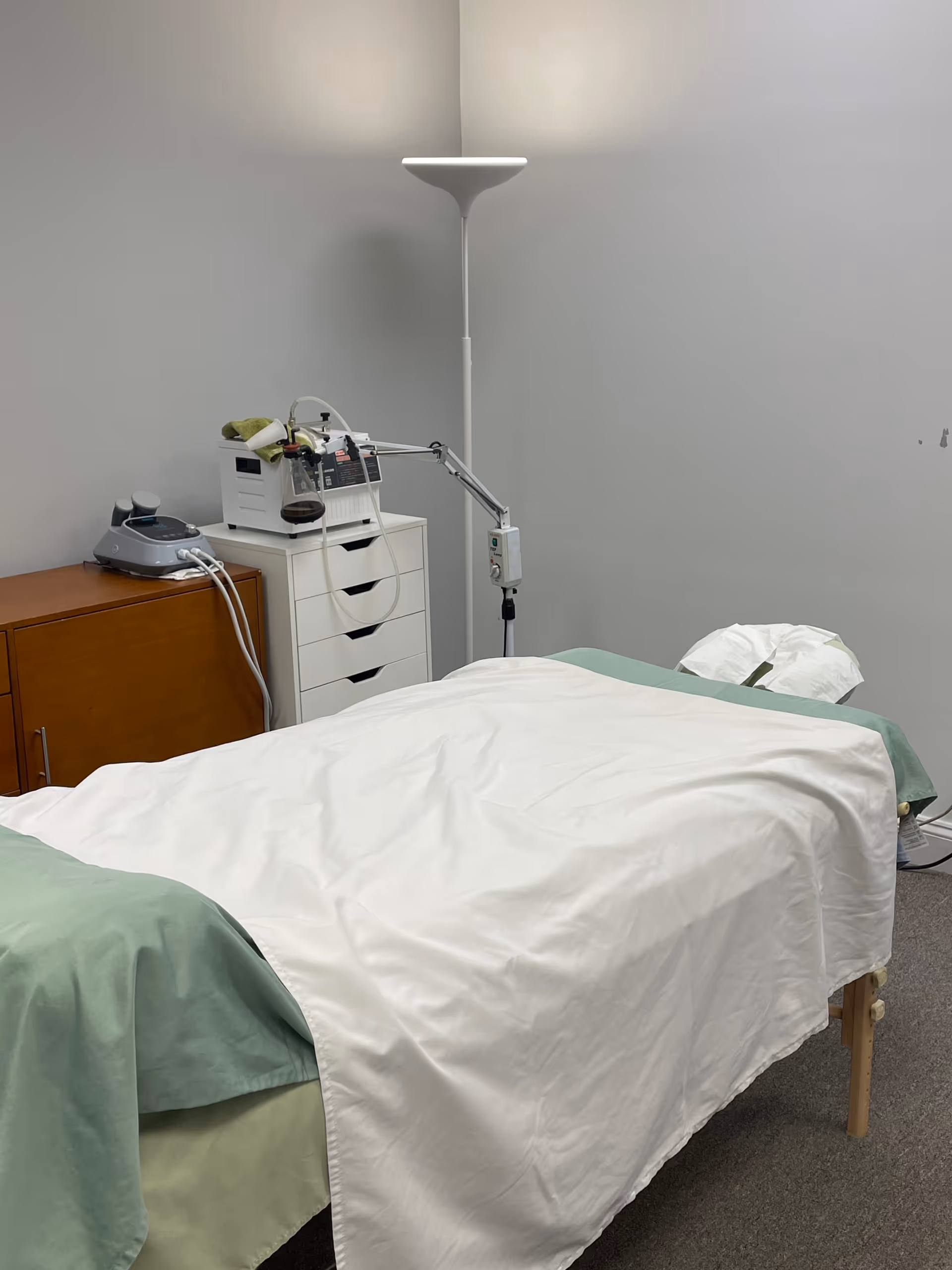 A clinical treatment room with a bed, medical equipment, and a tall floor lamp.