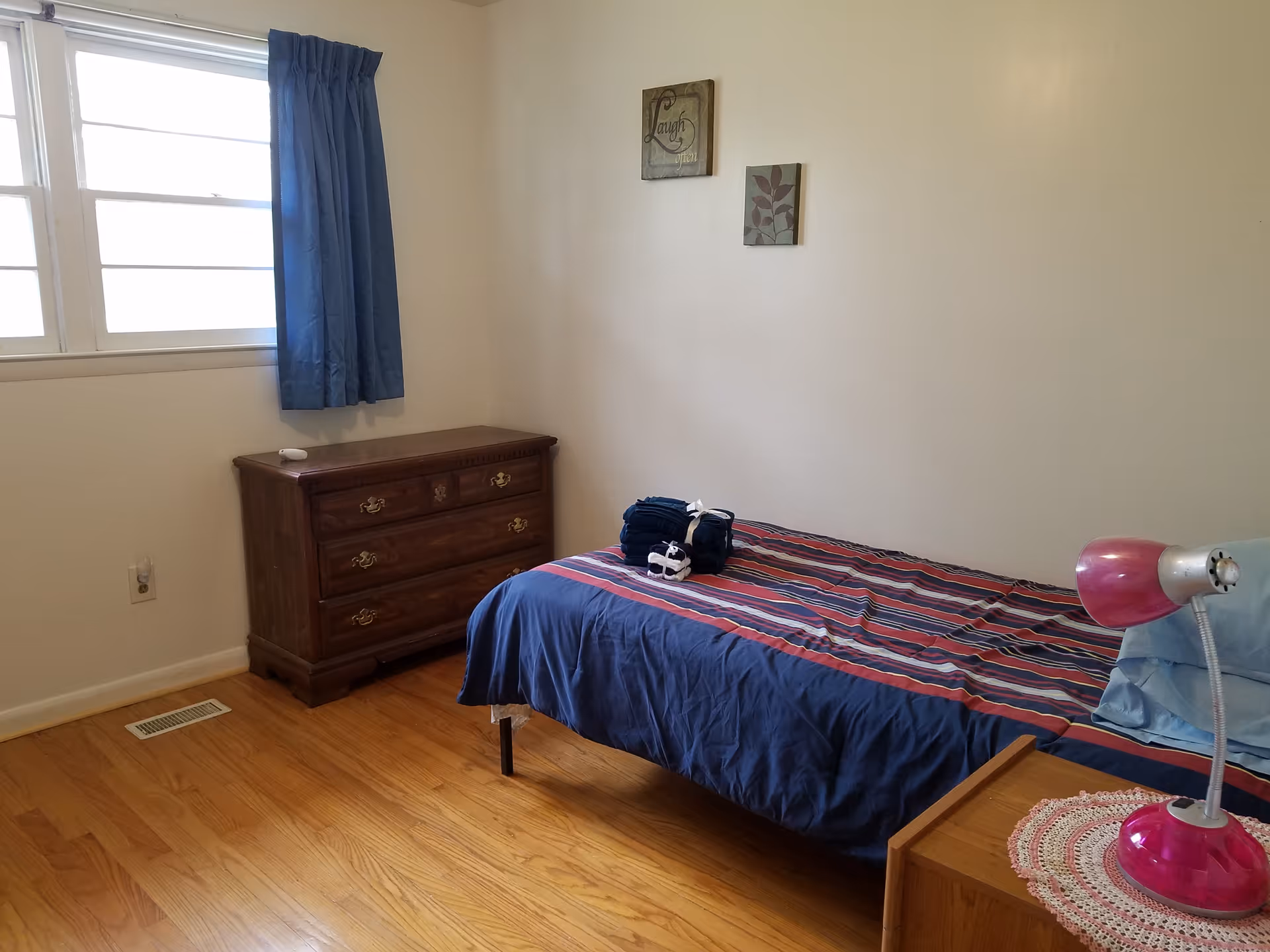 Simple furnished bedroom with bed, dresser, window, striped bedding, and pink lamp.