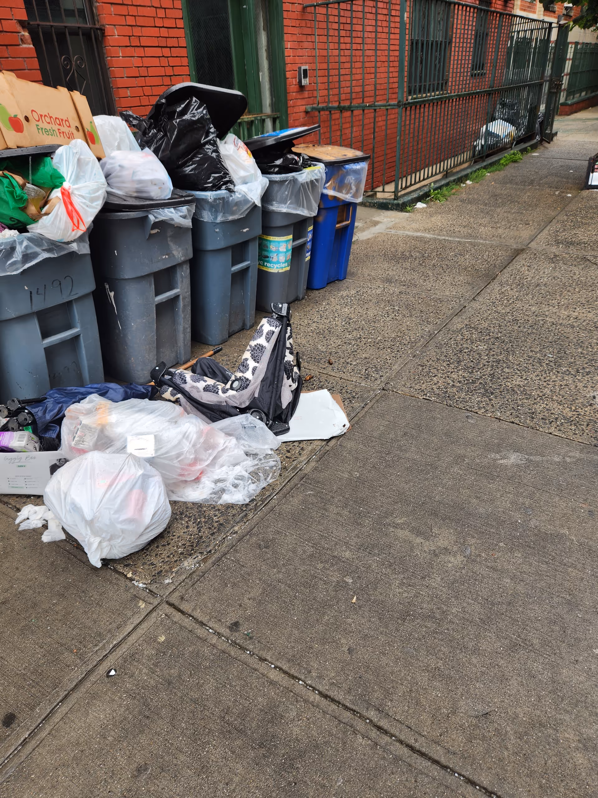 Trash bins, bags, and debris lining a city sidewalk beside a brick building.