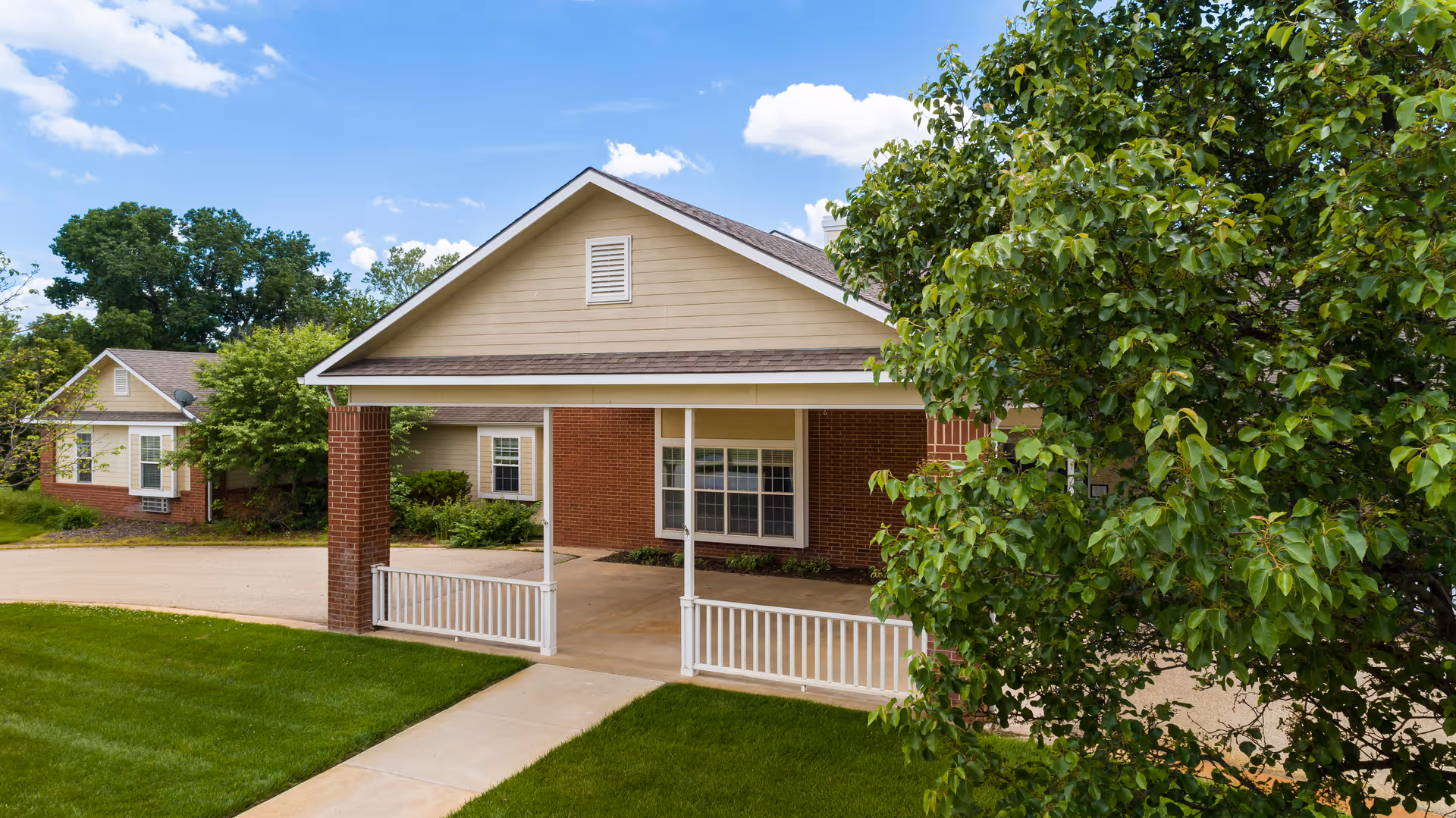 Front entrance of a single-story brick assisted living building with a covered driveway and surrounding trees and lawn.